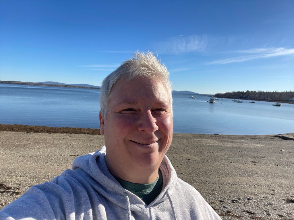 Author standing on rocky beach with calm waters on autumn, sunny day.